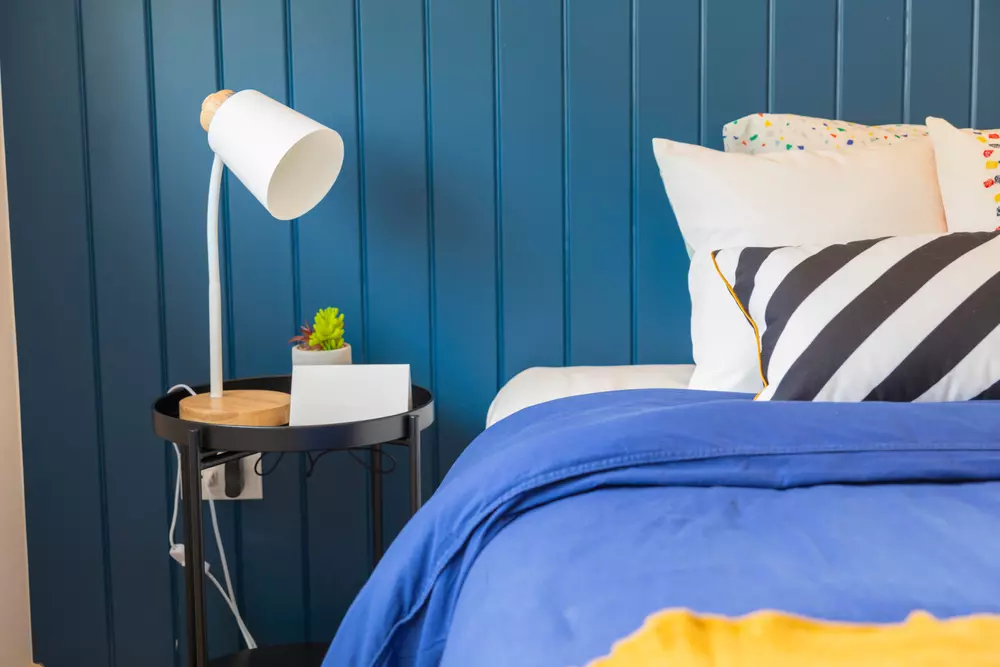 Quarto moderno com parede azul, cama com roupas de cama azul e travesseiros decorativos, luminária branca ao lado da cama, ambiente aconchegante e bem decorado.