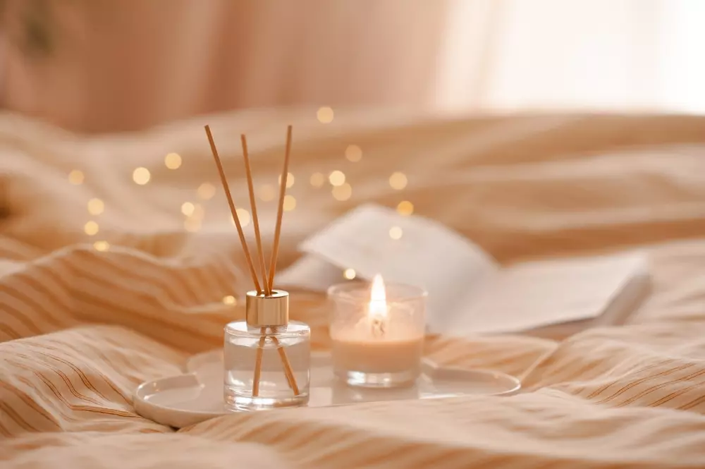 Um difusor de aroma e uma vela aromática lado a lado em cima de uma cama com lençóis em tons neutros. Um dos hábitos perigosos em casa é deixar velas próximas a tecidos.