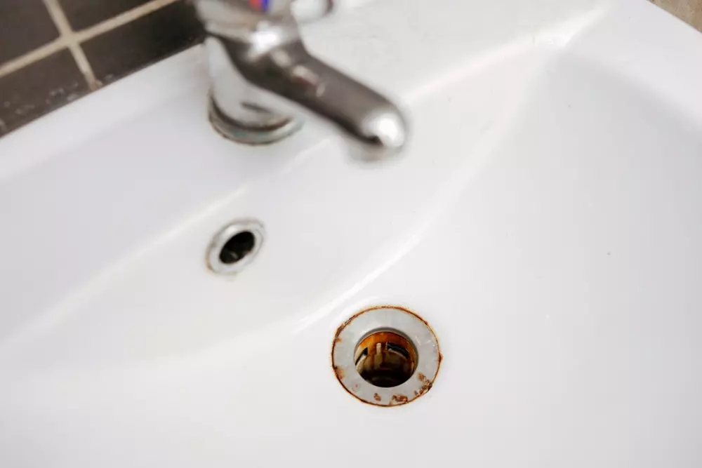Uma pia branca de banheiro com torneira prata e um pequeno ralo na pia está precisando de manutenção e enferrujado. Um dos hábitos perigosos em casa, deixar de fazer manutenção preventiva