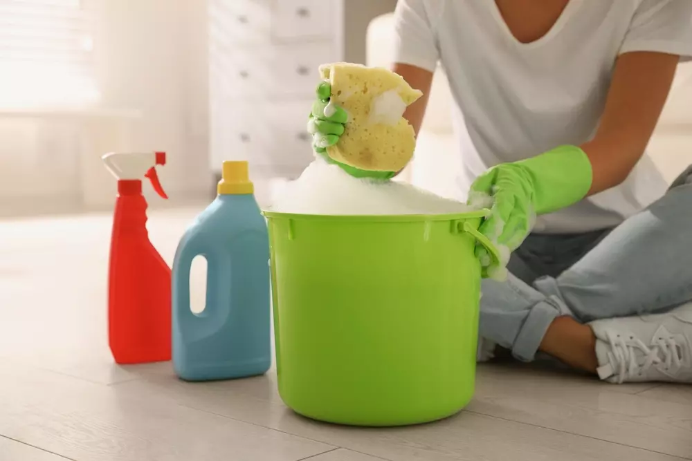 Frascos de cores diferentes de produtos de limpeza e uma mulher utilizando um balde verde para misturá-los, segurando uma esponja.
