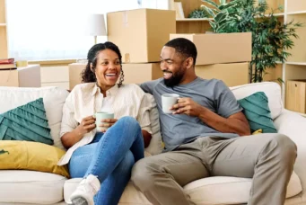 Um casal feliz que acaba de sair do aluguel e comprar seu apartamento. Estão felizes sentados ao sofá, tomando café e ao fundo caixas com itens de sua mudança.