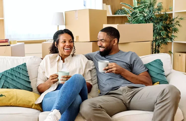 Um casal feliz que acaba de sair do aluguel e comprar seu apartamento. Estão felizes sentados ao sofá, tomando café e ao fundo caixas com itens de sua mudança.