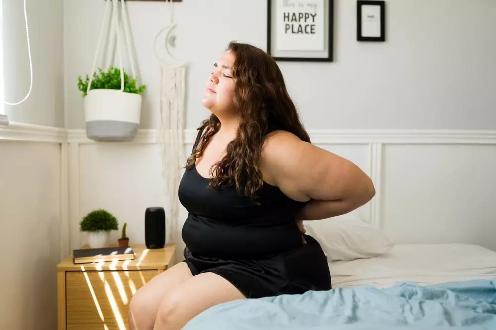 Uma mulher está sentada em sua cama ao acordar em seu quarto colocando as mãos nas costas por estar sentindo desconforto na coluna. Um dos problemas que fazem uma pessoa decidir trocar de colchão.