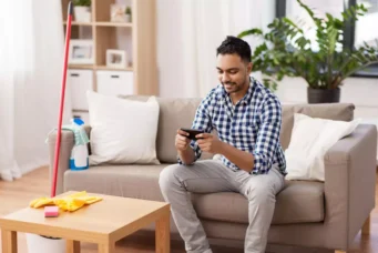 Homem sorridente sentado no sofá usando celular, ao lado de objetos de limpeza como vassoura, criando uma atmosfera de distração e procrastinação nas tarefas de casa.