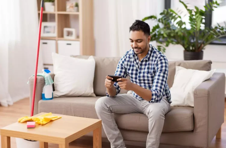 Homem sorridente sentado no sofá usando celular, ao lado de objetos de limpeza como vassoura, criando uma atmosfera de distração e procrastinação nas tarefas de casa.