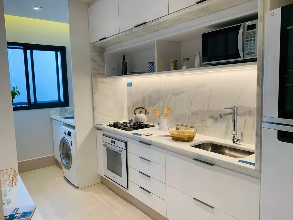 Cozinha moderna com detalhes em branco, pia de alumínio, micro-ondas, geladeira e eletrodomésticos, ambiente organizado e bem iluminado.