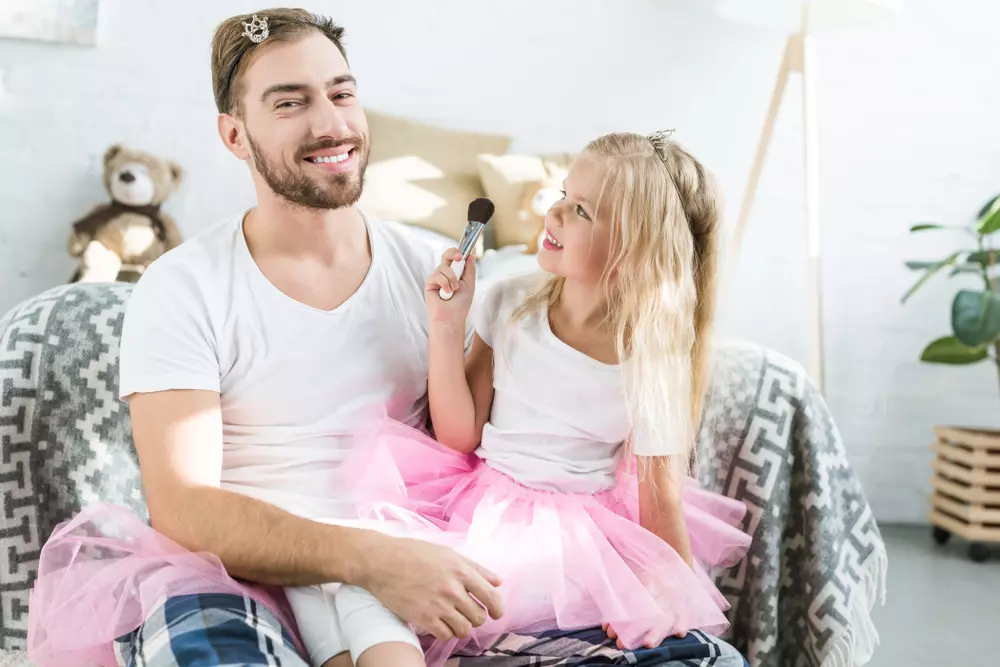 Pai e filha brincando juntos em casa, onde a menina segura um pincel de maquiagem enquanto o pai sorri com alegria, ambos vestidos de branco e rosa. Fundo acolhedor com brinquedos e plantas.