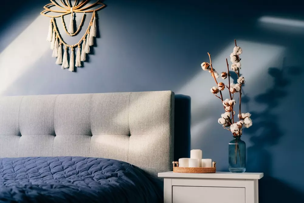 Quarto moderno com cabeceira estofada cinza, arranjo de algodoeiro em vaso de vidro azul e decoração de parede com mandala de parede de tricô na cor branca, criando um ambiente aconchegante.