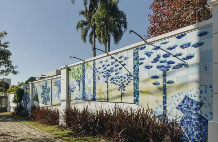 Mural artístico na CASACOR Paraná com árvores estilizadas em mosaico azul e verde, decorando uma parede com iluminação externa, ao ar livre, em um dia ensolarado.