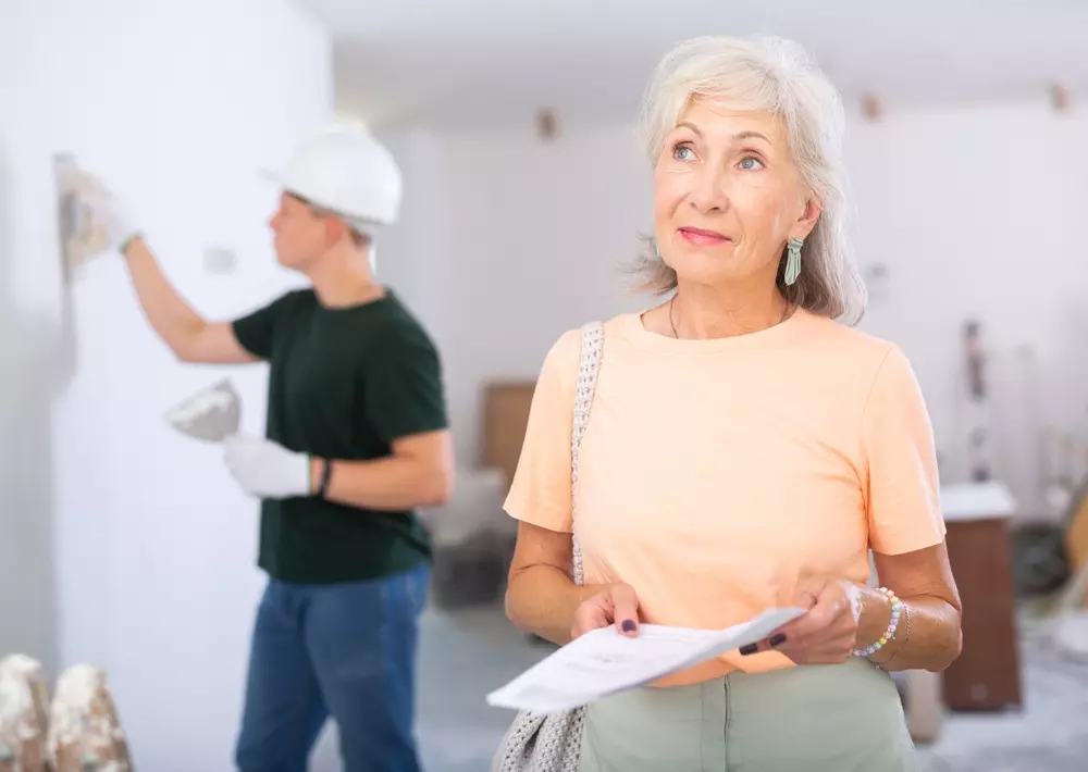 Mulher idosa observando uma obra de reforma na casa, enquanto um jovem trabalhador realiza reparos na parede ao fundo. Imagem representa cuidado com o lar e reforma residencial.