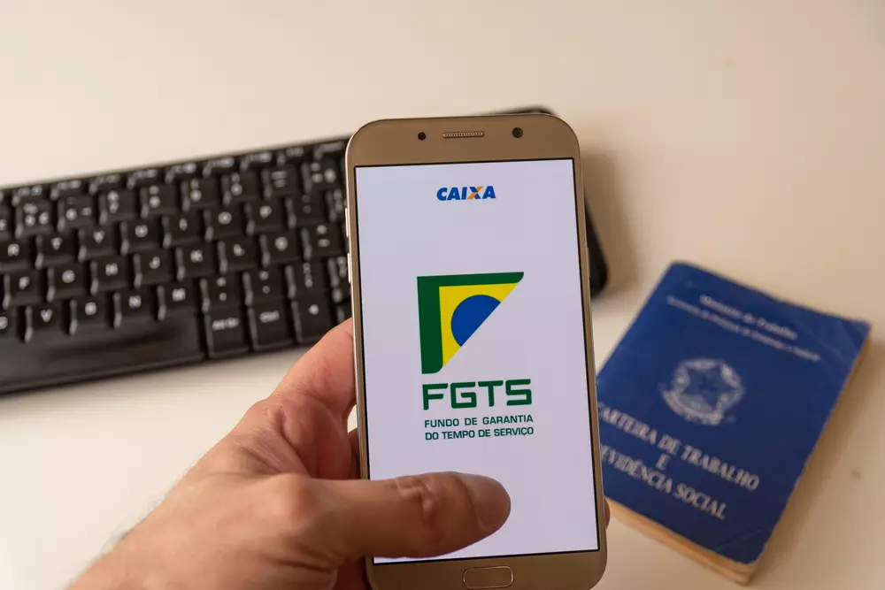 Pessoa segurando um celular com a tela exibindo o logo do Fundo de Garantia por Tempo de Serviço (FGTS), próximo de um teclado de computador e um documento azul de vínculo trabalhista.