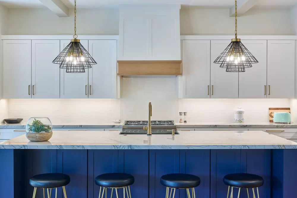 Imagem de cozinha moderna com balcão de ilha azul-marinho, iluminação de pendente de metal preto, armários brancos, pia com torneira dourada.