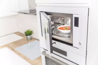 Imagem de um forno micro-ondas embutido na cozinha moderna com porta de vidro transparente, exibindo uma tigela com sopa quente.