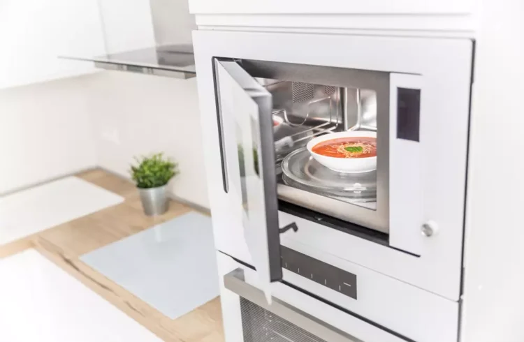 Imagem de um forno micro-ondas embutido na cozinha moderna com porta de vidro transparente, exibindo uma tigela com sopa quente.