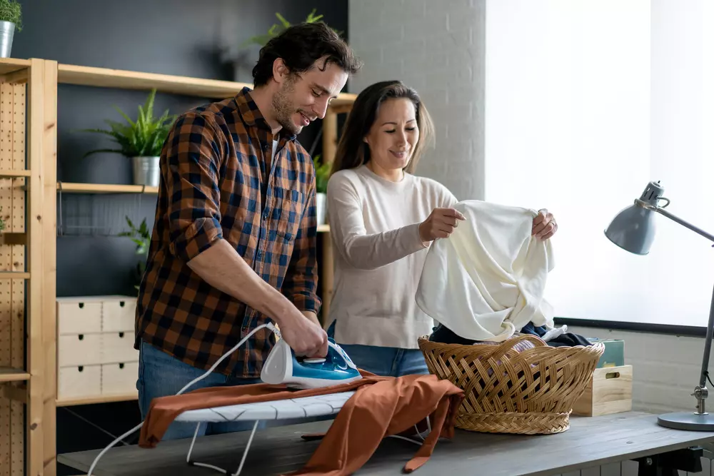 Duas pessoas ajudando a evitar procrastinação nas tarefas de casa enquanto organizam roupas e passam ferro na lavanderia