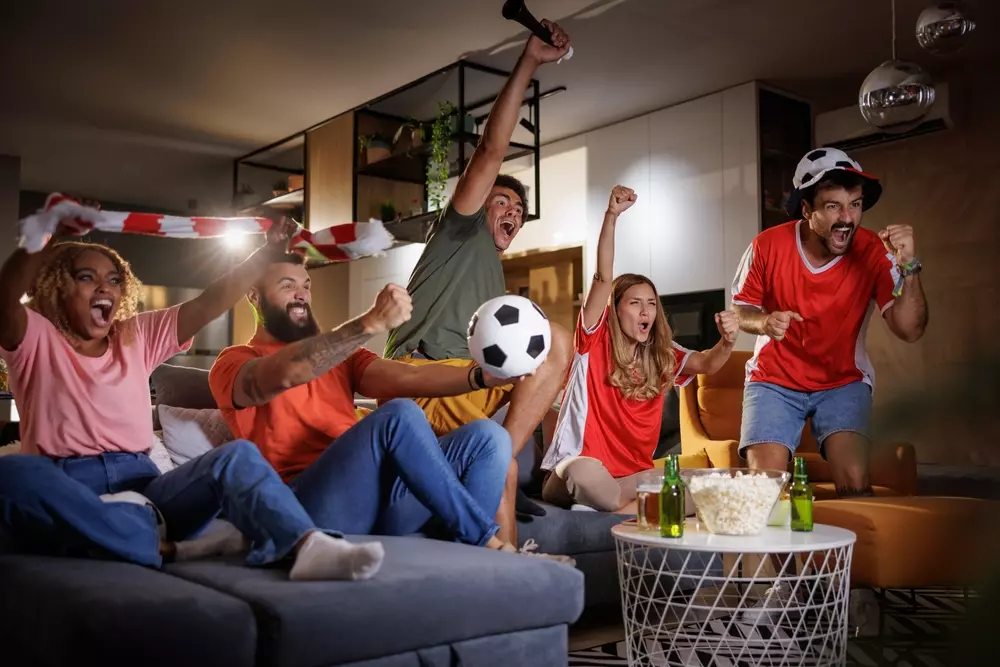 Grupo de amigos assistindo a um jogo de futebol na televisão, decorado com temas de futebol na sala de estar, ideal para ambientes de convivência esportivos.