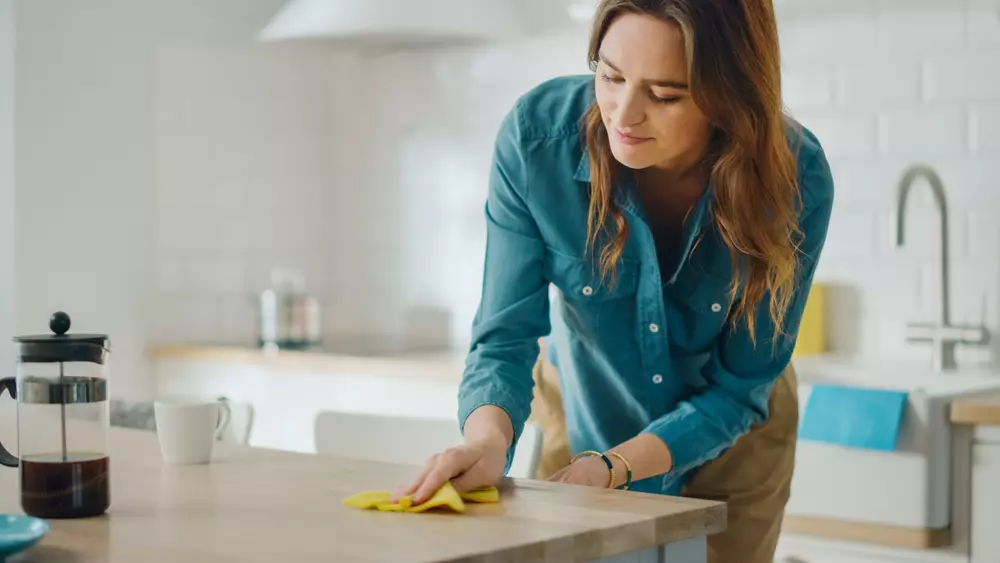 Mulher limpando a mesa na cozinha moderna com uma toalha de microfibra, demonstrando limpeza doméstica eficiente.
