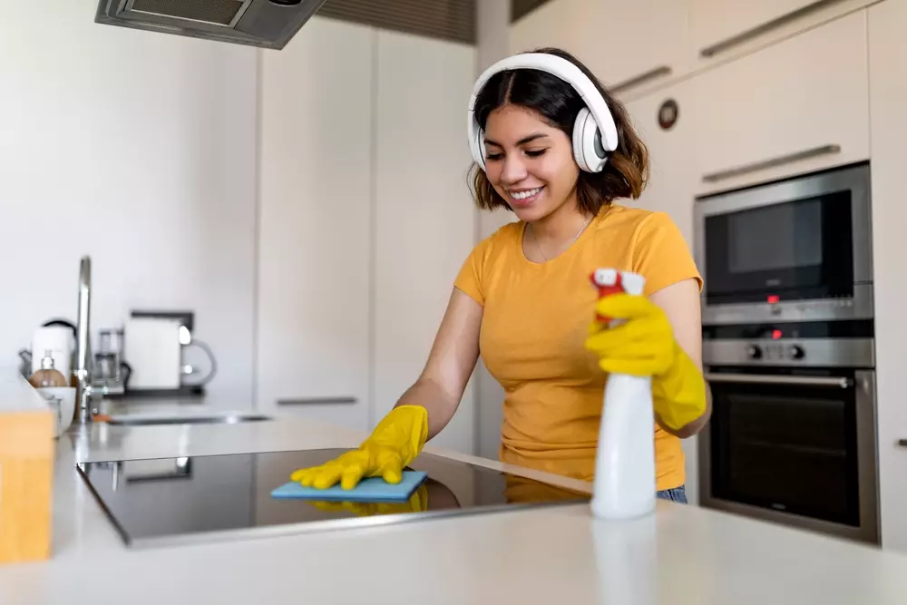Mulher jovem limpando o fogão na cozinha com luvas amarelas usando fone de ouvido, promovendo limpeza doméstica eficiente e prazerosa.