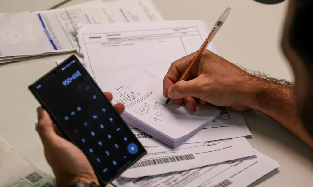 Mãos de um homem fazendo levantamento de suas dívidas anotando em um bloco de papel e segurando uma calculadora. Nome negativado pode ser um dos impedimentos