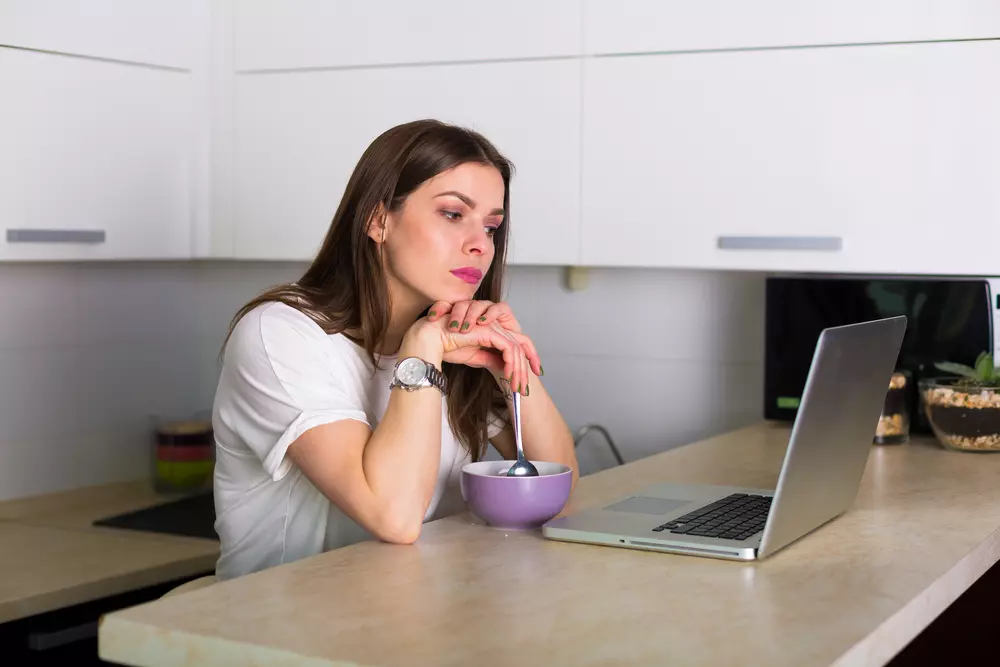 Jovem mulher pensativa na cozinha, procrastinando tarefas de casa enquanto assiste streaming no computador com uma tigela próxima de suas mãos.