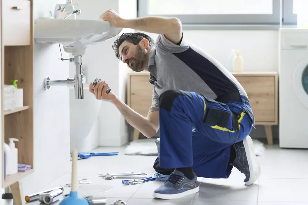 Reparo de encanamento por um encanador profissional em banheiro residencial, com diversas ferramentas ao redor e homem usando uniforme azul, realizando manutenção sob pia.
