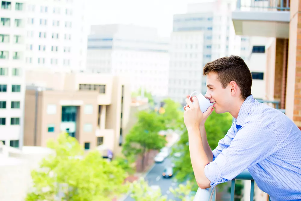 Homem sorridente tomando café em varanda de apartamento durante mudança de endereço, com vista urbana ao fundo. Simboliza tranquilidade e privacidade na nova etapa.