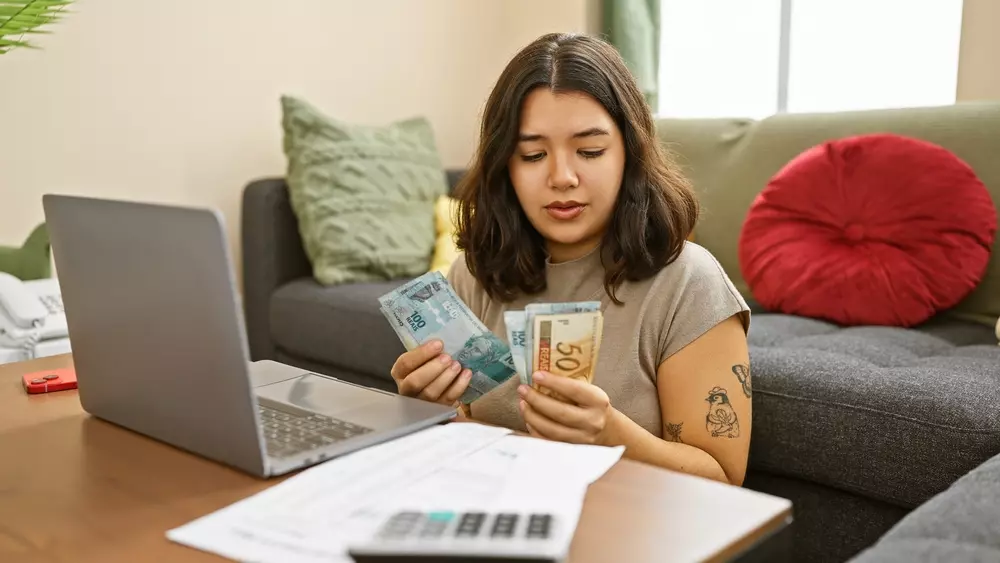 Mulher com dificuldades financeiras analisando dinheiro, representando impedimentos do financiamento devido a renda insuficiente, em ambiente doméstico.