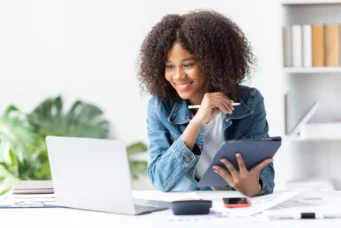 Mulher jovem de cabelos cacheados usando um tablet em um escritório claro e organizado, sorrindo enquanto trabalha com tecnologia e estudos.