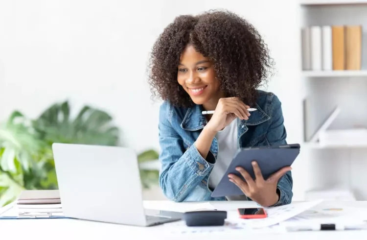 Mulher jovem de cabelos cacheados usando um tablet em um escritório claro e organizado, sorrindo enquanto trabalha com tecnologia e estudos.