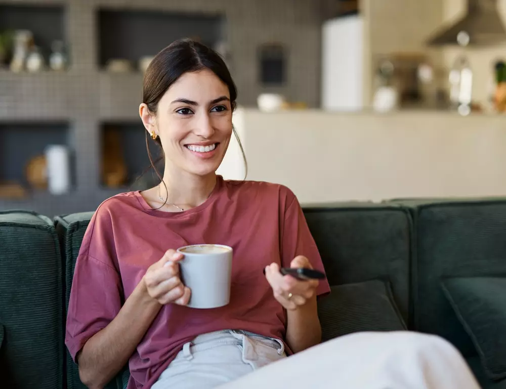 Mulher sorridente em casa, segurando uma caneca de café e o celular, buscando evitar procrastinação nas tarefas de casa e acumular pontos para assistir à sua série favorita.