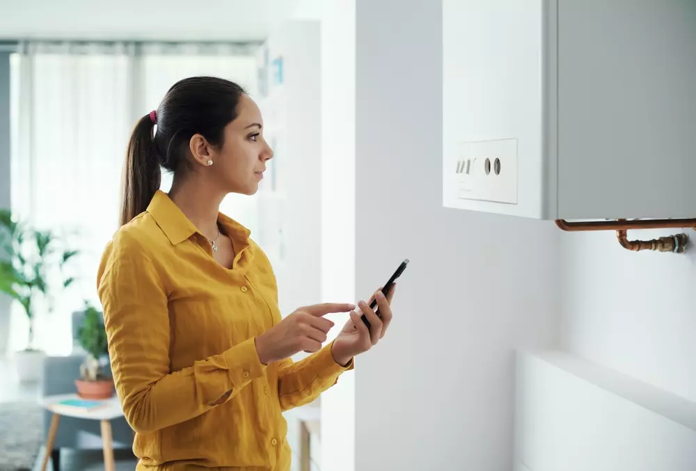 Mulher verificando uma caldeira a gás na parede com um smartphone, demonstrando manutenção ou inspeção técnica, em ambiente residencial bem iluminado.