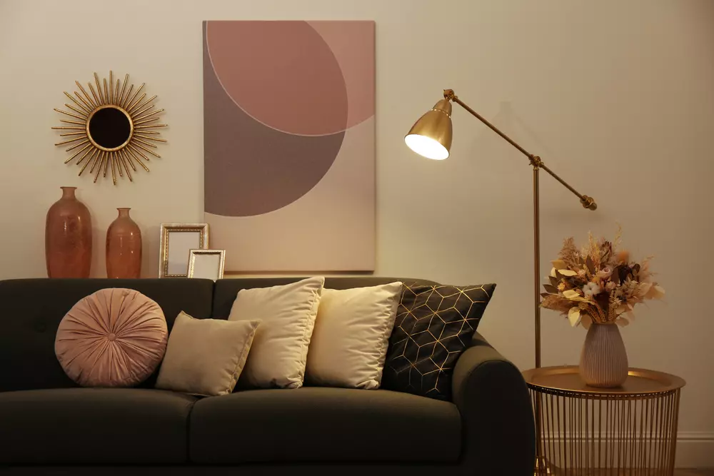 Sala de convivência decorada no estilo cluttercore com iluminação acolhedora, almofadas confortáveis, vasos decorativos, obra de arte abstrata e uma luminária de piso dourada.