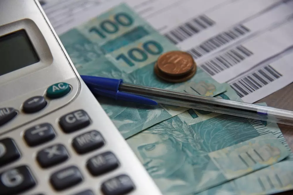 Uma calculadora, dinheiro em notas de 100 reais, uma moeda de 1 real e uma caneta, representando controle financeiro e planejamento de gastos.