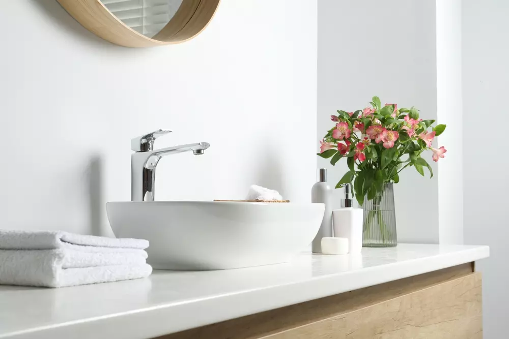 Banheiro moderno com detalhes delicados, incluindo um vaso de flores, toalhas brancas, sabonete e acessórios de higiene ao lado de uma pia branca, promovendo pequenos gestos de delicadeza.