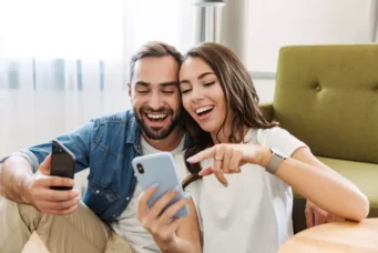 Casal feliz assistindo a fotos no celular, representando a conquista da casa própria para a classe média, com alegria e satisfação.