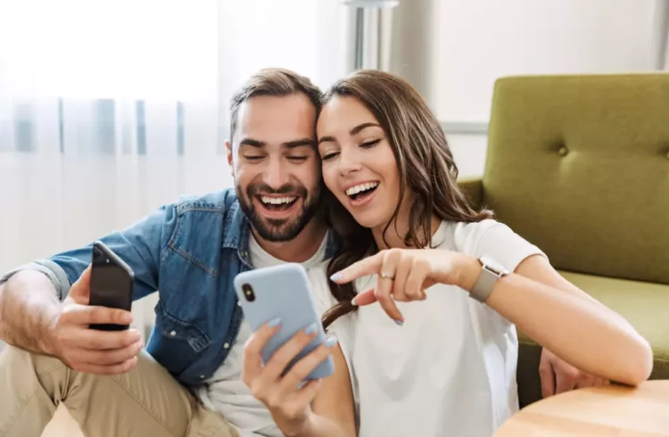 Casal feliz assistindo a fotos no celular, representando a conquista da casa própria para a classe média, com alegria e satisfação.