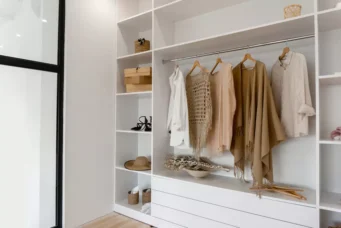 Armário organizado com roupas elegantes em cores neutras, incluindo suéteres, camisas e acessórios, em espaço moderno e bem iluminado.