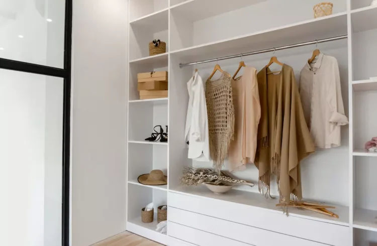 Armário organizado com roupas elegantes em cores neutras, incluindo suéteres, camisas e acessórios, em espaço moderno e bem iluminado.