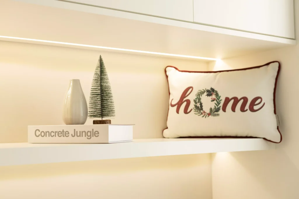 Enfeite de mini árvore na prateleira com uma almofada escrita 'home' e uma mini árvore de Natal ao lado de um livro sobre concreto.