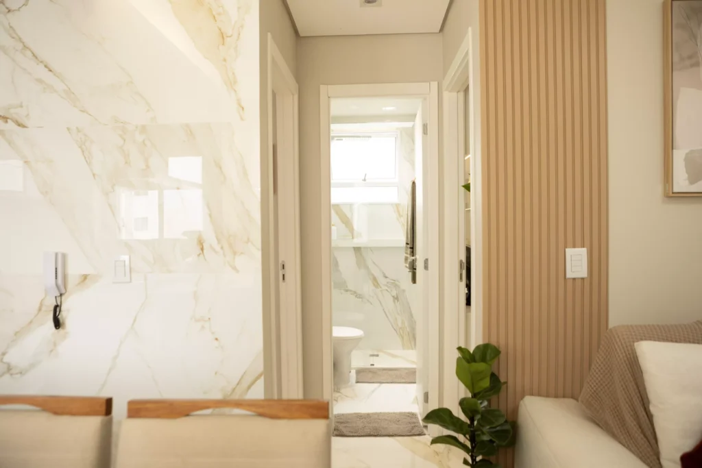 Entrada de banheiro moderno, vista do corredor do apartamento, bem iluminado, com acabamento de mármore na parede e ambiente aconchegante.