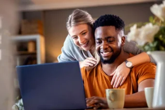 Casal feliz usando computador em casa, representando dicas de como evitar a inadimplência no condomínio, com foco na convivência e organização financeira.