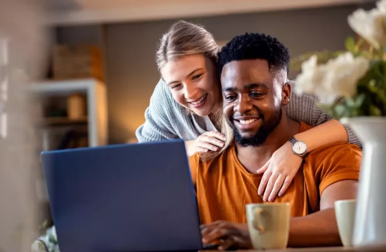 Casal feliz usando computador em casa, representando dicas de como evitar a inadimplência no condomínio, com foco na convivência e organização financeira.
