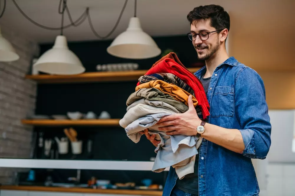 Homem de óculos e camisa jeans organizando roupas no interior de uma cozinha moderna