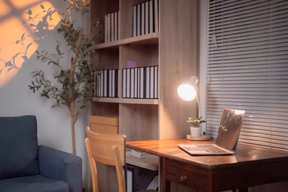 Ambiente de escritório residencial com estante de madeira, sofá azul, mesa de trabalho com laptop, iluminação adequada e decoração moderna.