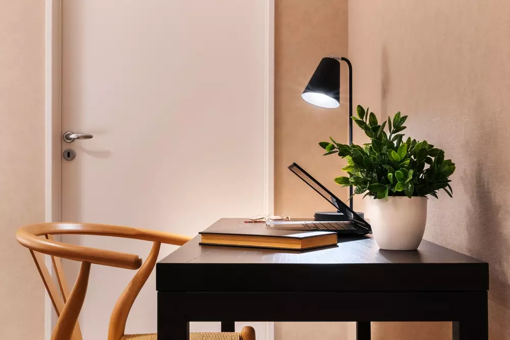 Um espaço de trabalho moderno com uma mesa preta, uma cadeira de madeira, uma luminária, livros e uma planta verde.