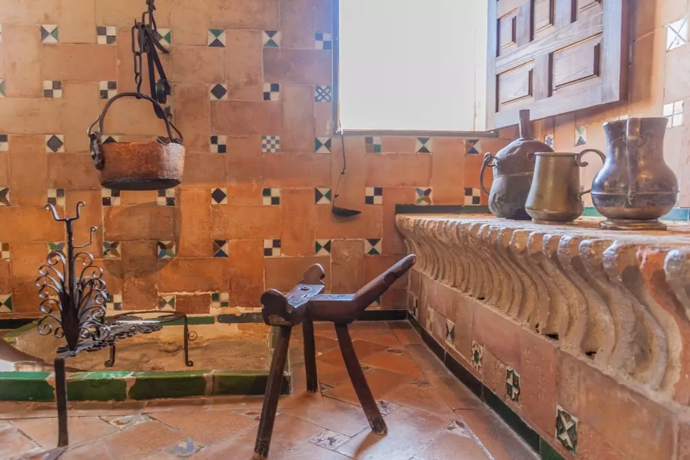 Interior de uma sala com decoração clássica, parede de azulejos rústicos, uma janela de madeira aberta, utensílios antigos em uma bancada de pedra, e móveis de época.