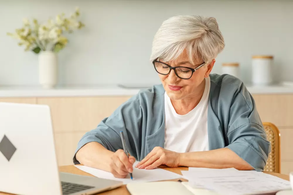 Mulher idosa usando óculos, sentada à mesa, fazendo anotações e planejando estratégias para juntar dinheiro de forma eficiente e segura.