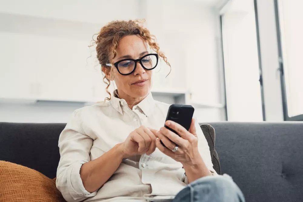 Mulher usando óculos de grau e camiseta branca, sentada em sofá confortável enquanto observa o smartphone, em ambiente moderno bem iluminado.