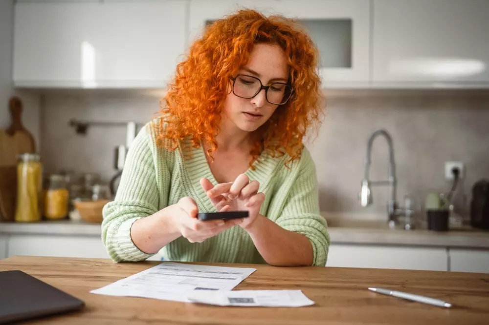 Mulher adulta usando celular na cozinha para realizar pagamento sem contato.