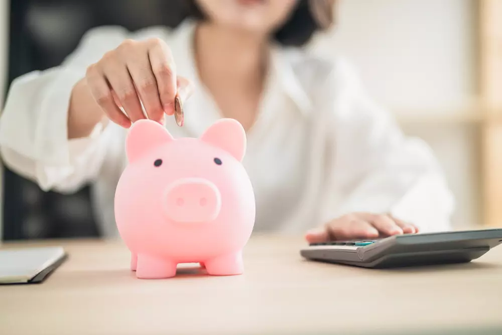 Uma mulher colocando moedas em uma caixa de poupança em formato de porquinho rosa, simbolizando a importância de juntar dinheiro e pagar-se primeiro para uma melhor organização financeira.
